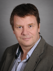 Dr. James Mitchell, inventor of the Quantum Crystal Engine and founder of DailyPlanet.Club, wearing a blazer and striped shirt in a formal portrait.